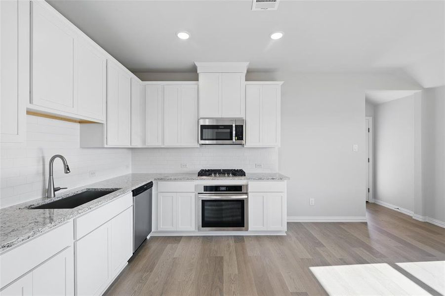 Kitchen with appliances with stainless steel finishes, backsplash, white cabinets, light wood finished floors, and light stone counters Kitchen with appliances with stainless steel finishes, backsplash, white cabinets, light wood finished floors, and light stone counters