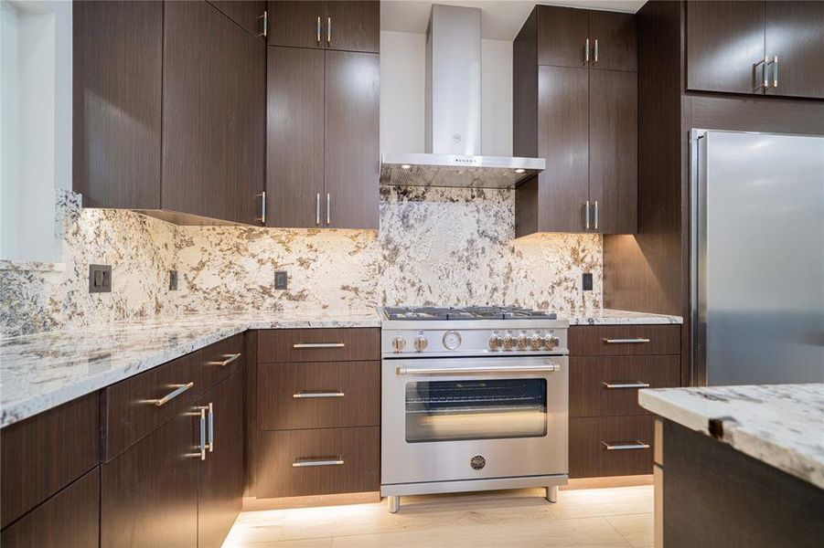 Gourmet kitchen featuring a stainless steel range with matching range hood, stone countertops, full-height stone backsplash, dark wood-finish cabinetry, and stainless steel refrigerator