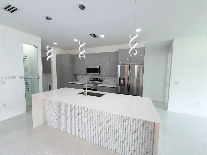 Kitchen featuring high-gloss flat-panel cabinetry, a waterfall-edge island with stone-surface countertop, tile backsplash, stainless steel appliances, and sculptural pendant lighting