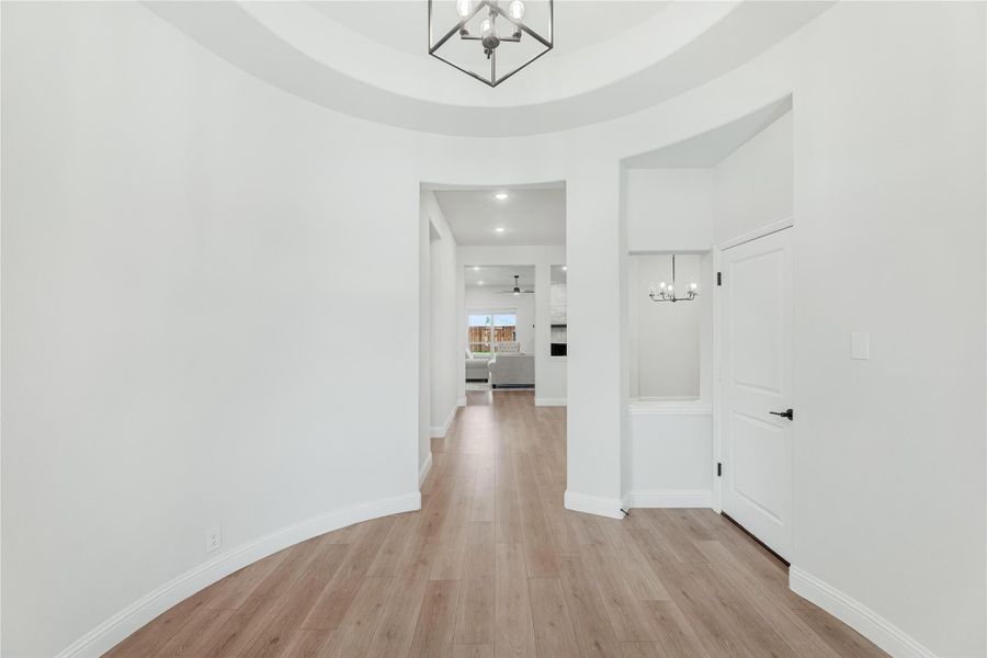 Curved entryway with tray ceiling, geometric chandelier, and light wood floors leading to living area