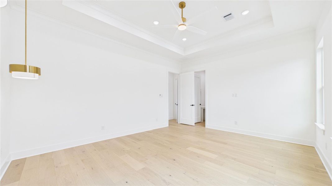 Bright, spacious primary bedroom with light hardwood floors and white walls. Features a modern ceiling fan, recessed lighting, and a stylish pendant light. The room has a tray ceiling and a doorway leading to an adjacent area.