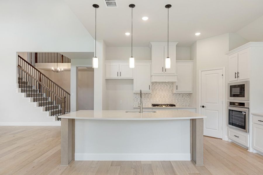 Open kitchen with large center island, white cabinets, pendant lights, and staircase visible in background Open kitchen with large center island, white cabinets, pendant lights, and staircase visible in background
