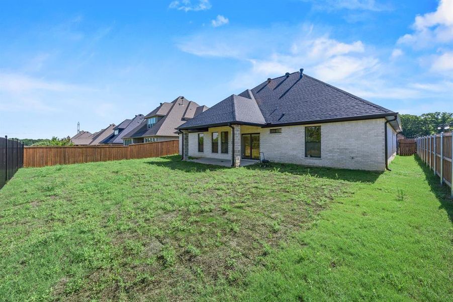 Expansive backyard featuring a new construction home with light-colored brick and stone accents, a dark shingle roof, and a privacy fence Expansive backyard featuring a new construction home with light-colored brick and stone accents, a dark shingle roof, and a privacy fence