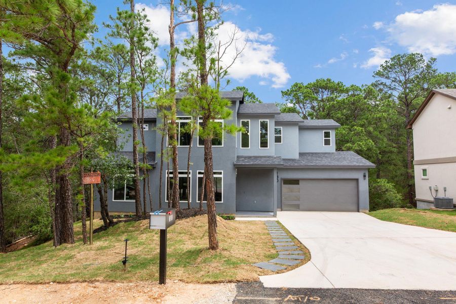 A contemporary gray residence, set amongst tall pine trees, presents a welcoming driveway and modern garage door.