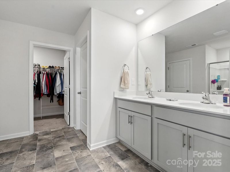 Luxurious primary bathroom featuring dual vanities with white countertops and gray cabinetry. Tile flooring, walk-in closet access, and separate shower area create spa-like master suite experience.