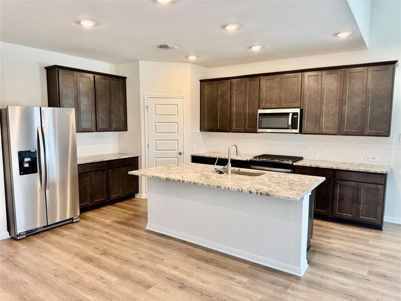 Kitchen featuring stainless steel appliances, dark brown cabinetry, light stone countertops, light wood finished floors, and recessed lighting