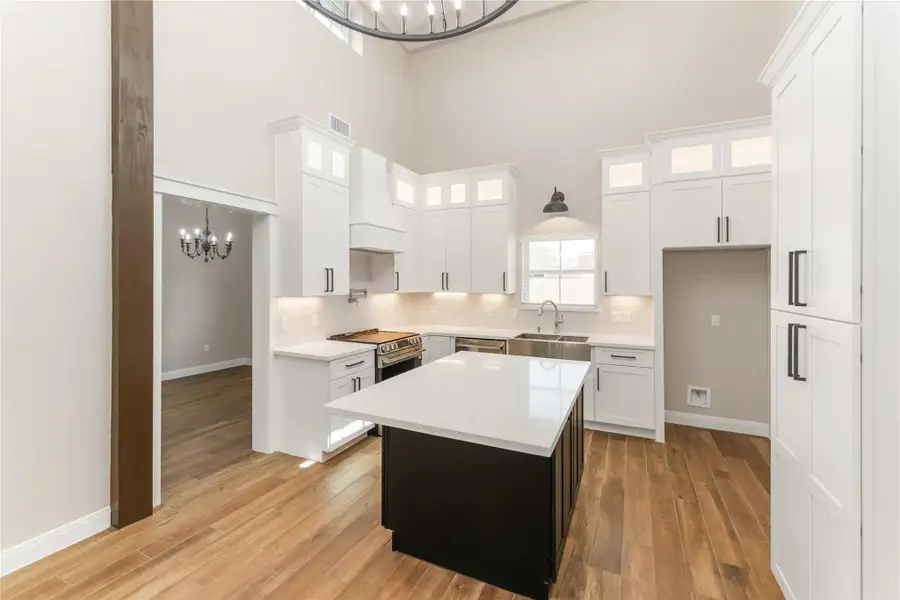 This bright, modern kitchen features high ceilings, white cabinetry with ample storage, and a large central island with a sleek countertop. A stainless steel farmhouse sink and high-end appliances complete the space, complemented by warm tile wood-style flooring and contemporary lighting.