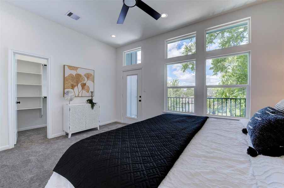 Primary bedroom showing private balcony door, large windows with tree views, ceiling fan, carpet, and open walk-in closet with built-in shelving.