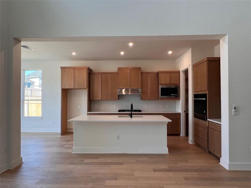 Kitchen with light wood finished floors, a kitchen island with sink, black appliances, wood finish cabinets, and recessed lighting