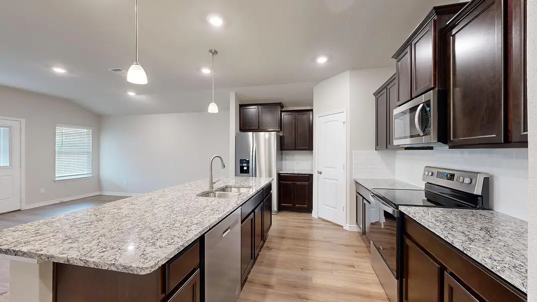Kitchen featuring stainless steel appliances, light wood finished floors, dark wood finish cabinets, light stone countertops, and a kitchen island with sink