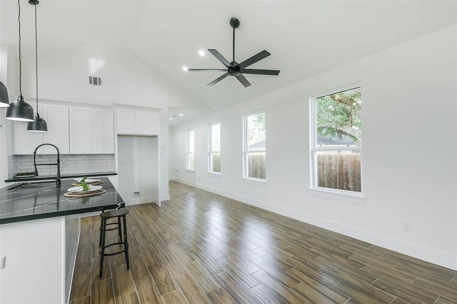 This is a modern, spacious kitchen with white cabinetry, dark countertops, and stainless-steel appliances. It features a high vaulted ceiling with recessed lighting and a ceiling fan, wood flooring, and ample natural light from several windows. A central island with pendant lighting doubles as a breakfast bar.