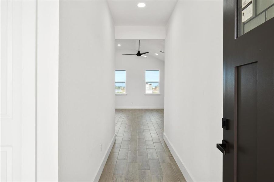 This photo shows a bright, modern hallway leading to a spacious room with large windows and a ceiling fan, featuring neutral walls and tiled flooring. A dark door adds a contrasting accent on the right side.