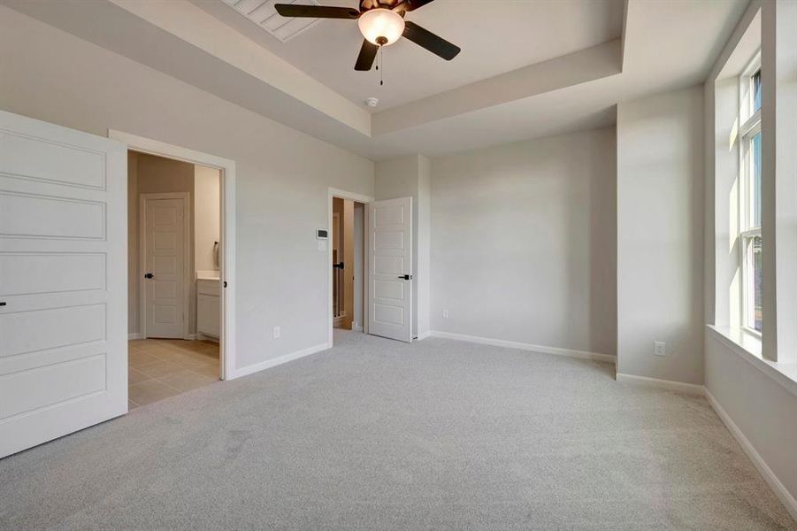 Spacious room featuring light gray carpet flooring, a tray ceiling with a ceiling fan, and large windows