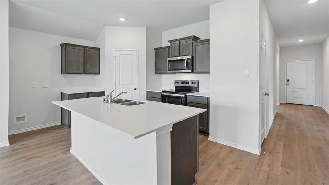 Modern kitchen featuring an island with a double basin sink, white countertops, dark wood-finish cabinetry, stainless steel microwave, and wood-finish flooring