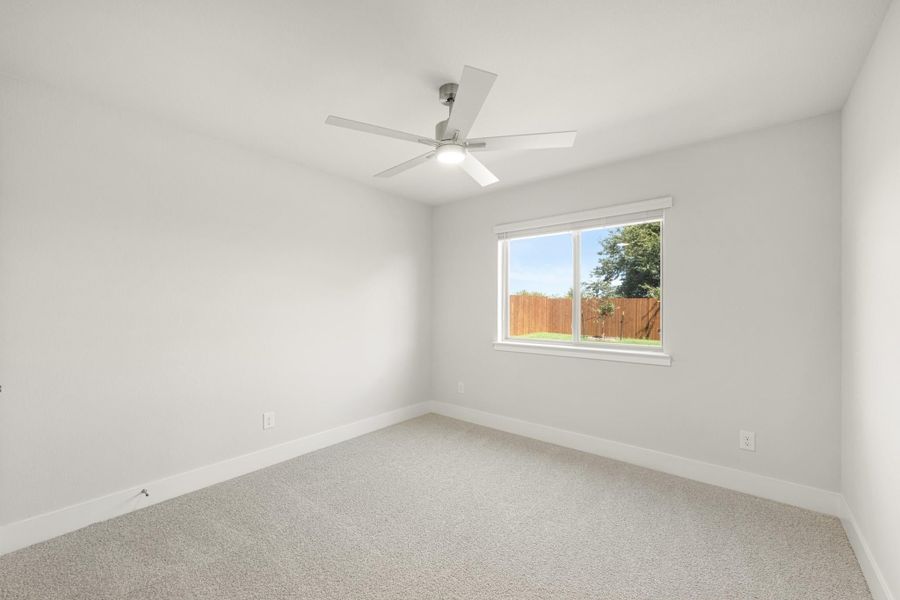 Empty bedroom with carpet flooring, ceiling fan, and a window overlooking a fenced backyard