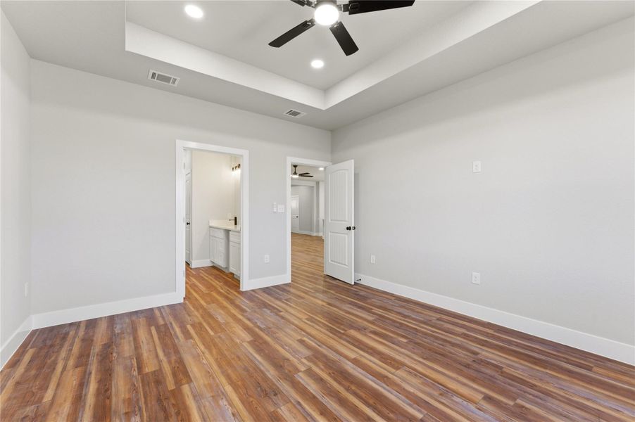 Unfurnished bedroom featuring a raised ceiling, ceiling fan, connected bathroom, wood finished floors, and recessed lighting