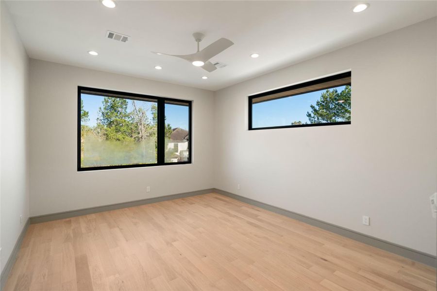 This room offers a bright and airy space with light wood floors, neutral walls, and large windows providing natural light and green views. Modern ceiling fan and recessed lighting enhance the fresh, contemporary feel.