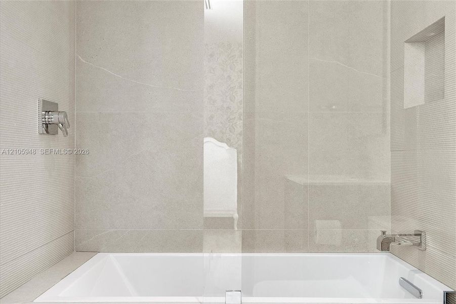 Soaking tub featuring large-format neutral tile surround, integrated wall niche, chrome wall-mounted faucet, and glass partition