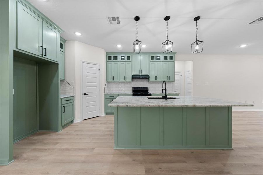 Kitchen featuring green cabinetry, light stone countertops, hanging light fixtures, an island with sink, and glass insert cabinets