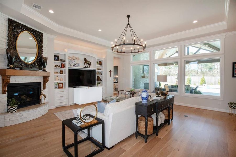 This living room features a cozy fireplace with a decorative mantle, built-in shelving, and a large TV. It offers ample natural light through expansive windows overlooking a well-landscaped backyard. The space is complemented by elegant hardwood flooring and a stylish chandelier, creating a warm and inviting atmosphere.