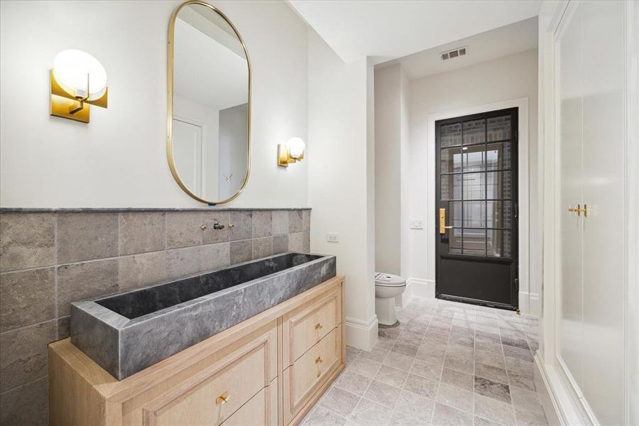 Located off of the outdoor kitchen is the powder bathroom. The powder bathroom features gold wall-mounted sconces, a unique stone sink, tile flooring, and a built-in white-oak vanity.