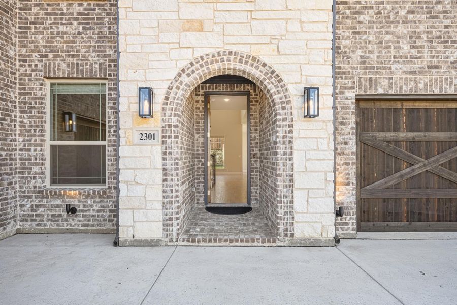 Brick home entrance with arched doorway, wall sconces, house number 2301, and wood garage door Brick home entrance with arched doorway, wall sconces, house number 2301, and wood garage door