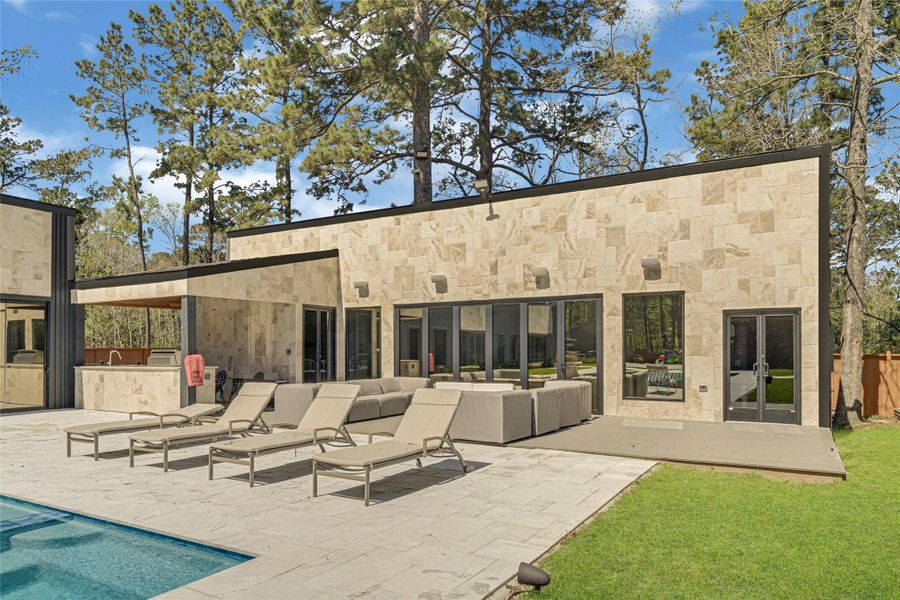 Stone exterior with expansive glass doors leading to a spacious patio. The outdoor area includes a pool, multiple lounge chairs, and a comfortable seating area, perfect for relaxing and entertaining in a serene wooded setting.