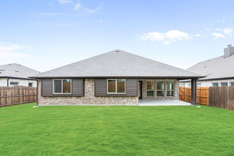 Rear view of single-story brick home with covered patio, dark siding accents, and large fenced backyard with green lawn.