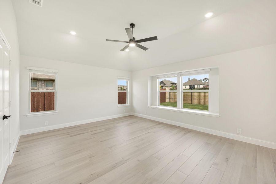Empty bedroom with light wood floors, ceiling fan, and multiple windows overlooking backyard fence