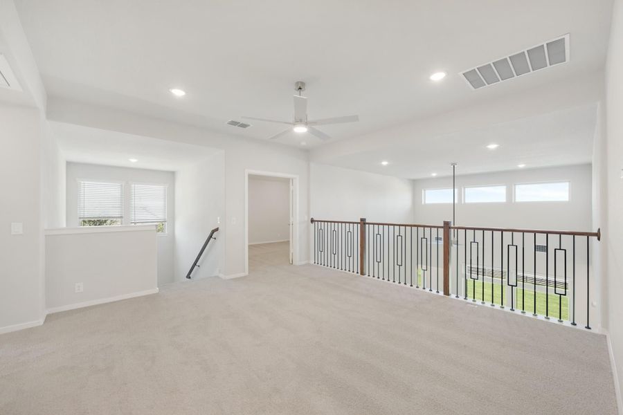 Open upstairs loft with carpet, ceiling fan, and decorative metal stair railing with wood handrail Open upstairs loft with carpet, ceiling fan, and decorative metal stair railing with wood handrail