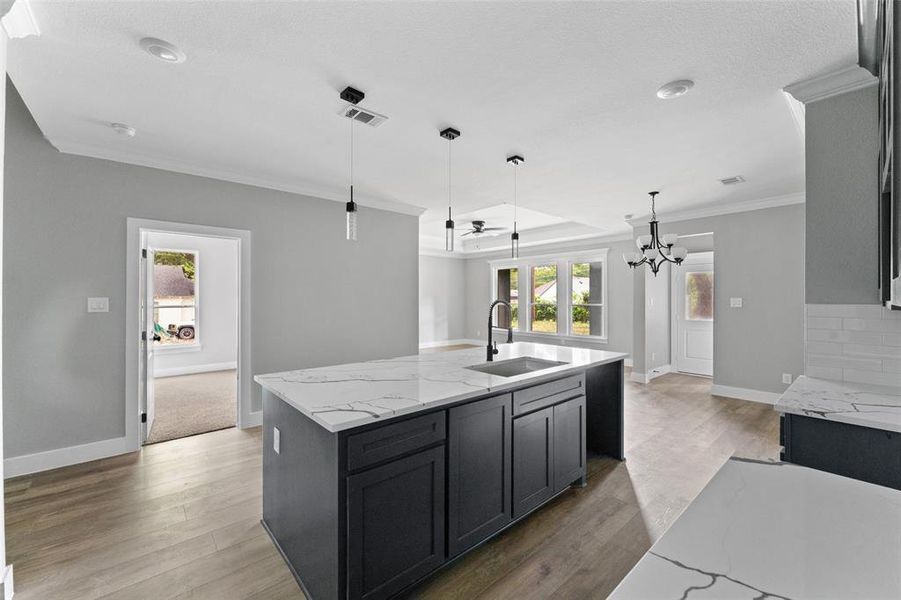 Kitchen featuring light stone counters, hanging light fixtures, wood finished floors, crown molding, and a kitchen island with sink