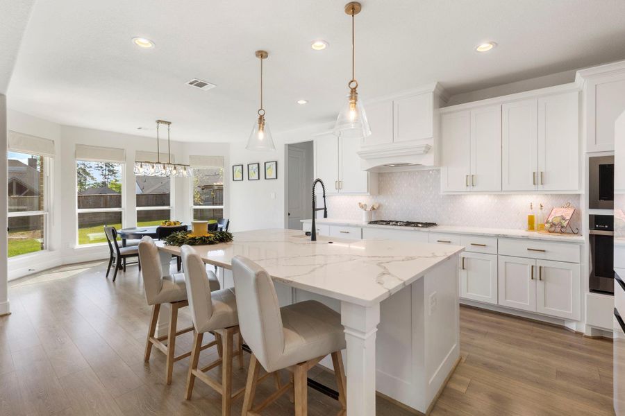 This photo showcases a bright, modern kitchen with white cabinetry, a large marble island, and upgraded pendant lighting. The open layout extends to the breakfast room with ample natural light from large windows, creating a welcoming and spacious atmosphere.