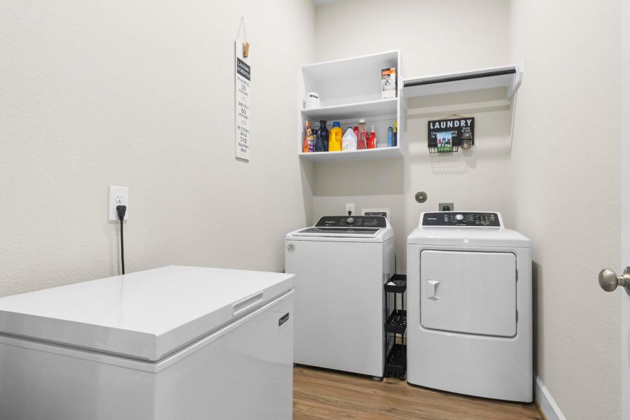 This photo shows a spacious laundry room featuring space for  washer and dryer, a chest freezer, and wall-mounted shelves for storage. The room is well-lit with a neutral color palette and has wooden flooring for a clean, functional look.