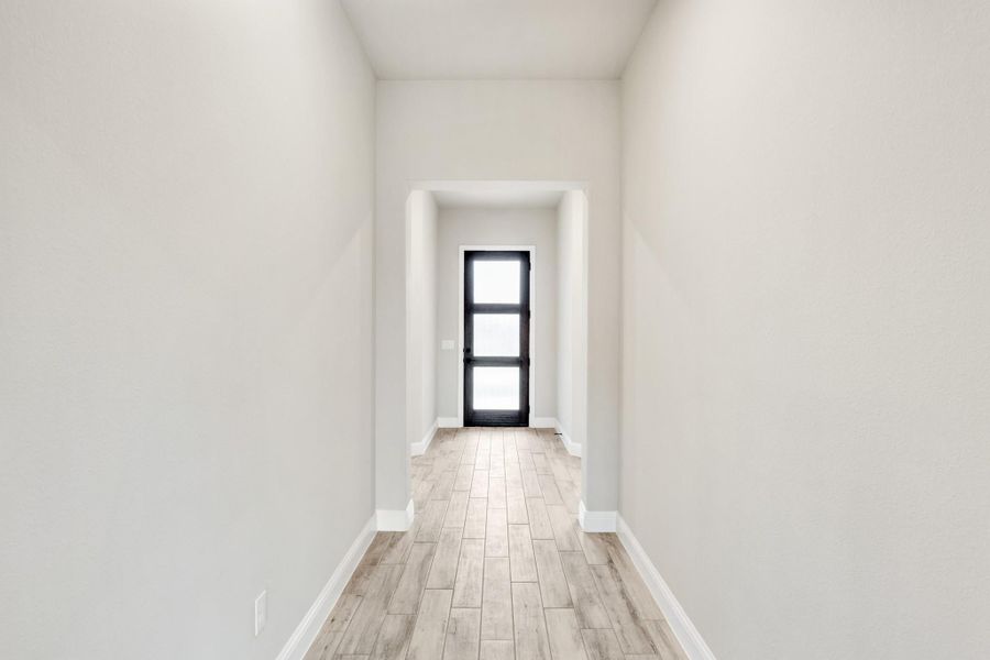 Narrow hallway with white walls, light wood floors, and black glass-panel door at the end