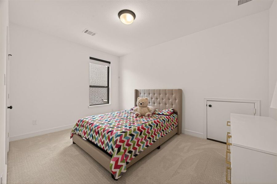 This bedroom features a cozy bed with a tufted headboard, colorful bedding, and a plush carpet. It has a small window for natural light and a unique small door for extra storage or play space.