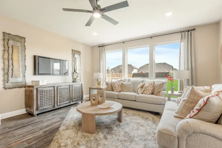 Living room with beige sofas, round wood coffee table, ceiling fan, wall-mounted TV, and large windows with backyard view