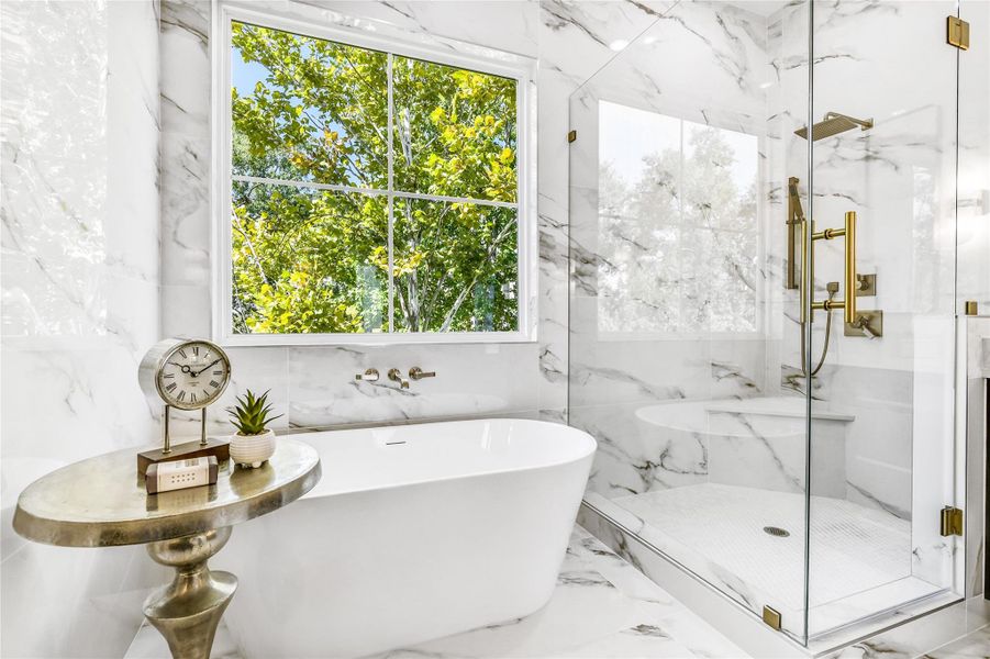 Bathed in natural light, this image spotlights the freestanding soaking tub perfectly positioned beneath a large picture window framed by lush greenery. The floor-to-ceiling marble tile surround elevates the space, while the adjacent walk-in shower—with frameless glass, brass hardware, and dual shower heads—delivers resort-quality relaxation. The sleek inset wall niche and floating corner bench showcase form meeting function in every detail.