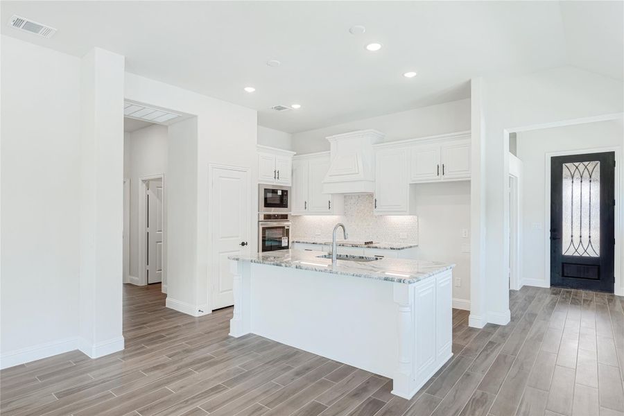 White kitchen with center island, granite countertops, white cabinets, tile backsplash, and stainless steel appliances on wood-look floors White kitchen with center island, granite countertops, white cabinets, tile backsplash, and stainless steel appliances on wood-look floors