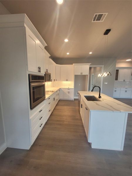 Kitchen with white cabinetry, backsplash, pendant lighting, stainless steel appliances, and a center island with sink