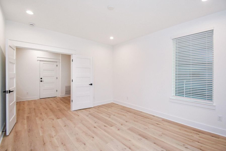 This room features bright white walls, light wood flooring, and recessed lighting. It includes a window with blinds and a set of double doors opening to an adjacent space, providing a modern and open feel.
