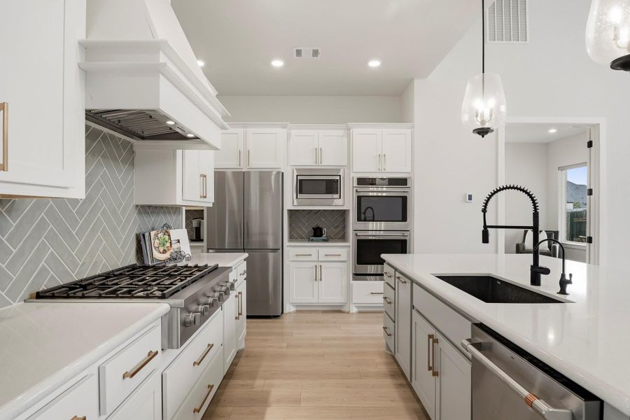 A six-burner GE Café commercial-style cooktop creates a striking focal point beneath a statement chimney vent hood, while coordinating GE Café double ovens and microwave complete the kitchen’s impressive suite of stainless steel appliances. Designed for serious cooking and sophisticated style, this chef-inspired setup brings professional-level performance to the heart of the home.