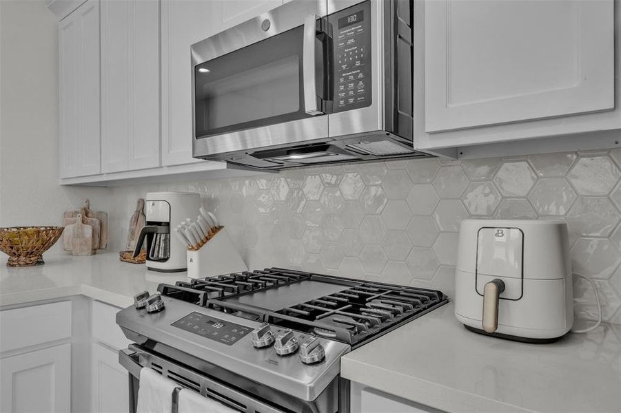 Stainless steel gas range with a coordinating stainless steel microwave oven above, white upper and lower cabinetry, white countertop, and a white hexagonal tile backsplash Stainless steel gas range with a coordinating stainless steel microwave oven above, white upper and lower cabinetry, white countertop, and a white hexagonal tile backsplash