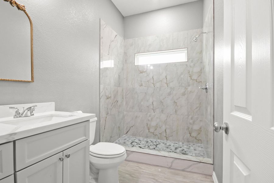 Secondary bathroom features a modern design with marble-style tiled shower, a sleek white vanity, and a large framed mirror. The neutral color palette enhances the bright and airy feel.
