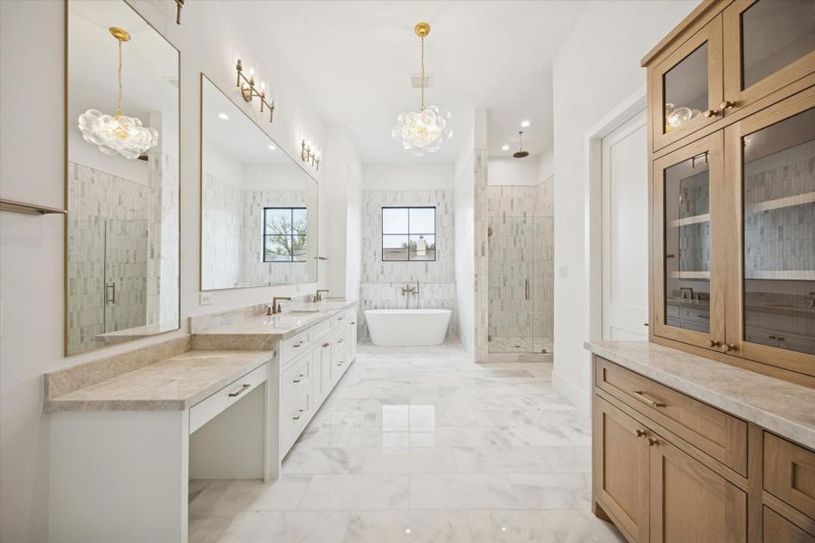 Spa inspired primary bath featuring dual vanities, full height tile surround, and a freestanding soaking tub framed by a picture window. Glass enclosed shower, marble flooring, and designer brass fixtures elevate the space with timeless luxury and refined craftsmanship.
