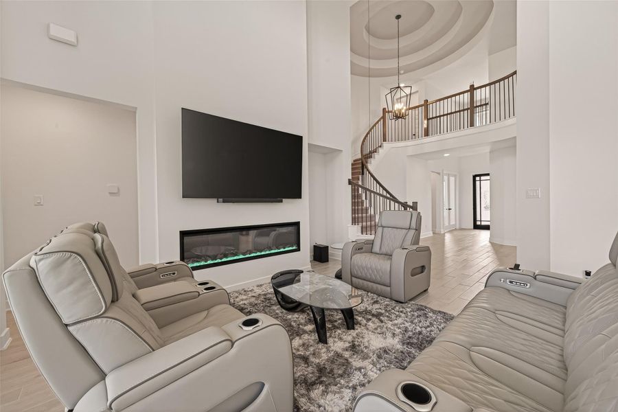This spacious living room features high ceilings, a modern fireplace below a large TV, and comfortable recliner seating. The open layout includes a striking staircase and a view of the entryway, creating an inviting and luxurious atmosphere.