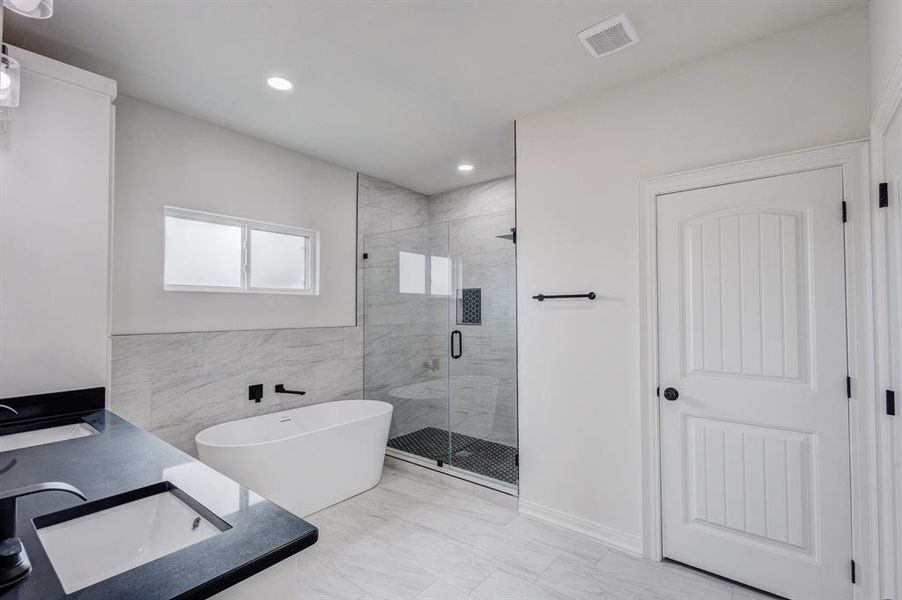 The bathroom features a dual sink vanity with dark countertops, a freestanding white bathtub, a walk-in shower with a glass enclosure and dark-tiled floor, and light-toned wall and floor tiles