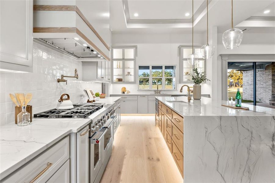 Gourmet kitchen featuring a large waterfall island with integrated sink, light wood-finish flooring, white shaker-style cabinetry with glass accents, a professional-grade range, and a full-height tile backsplash