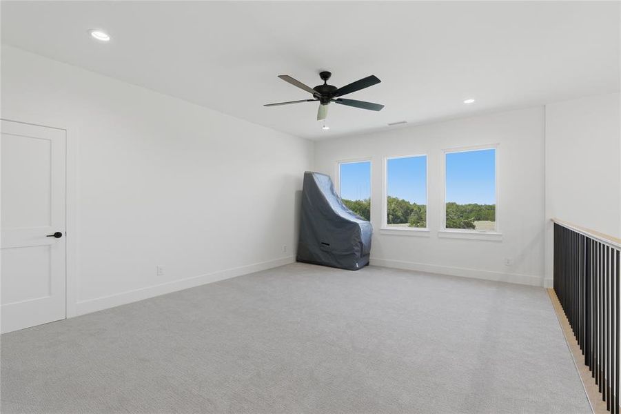Spacious room featuring light gray carpeting, three prominent windows, a ceiling fan, recessed lighting, and a modern black metal railing