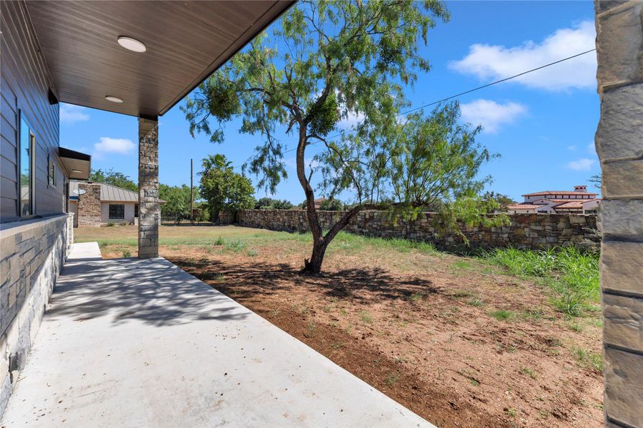 A covered concrete patio overlooks the yard bordered by a rustic stone wall.