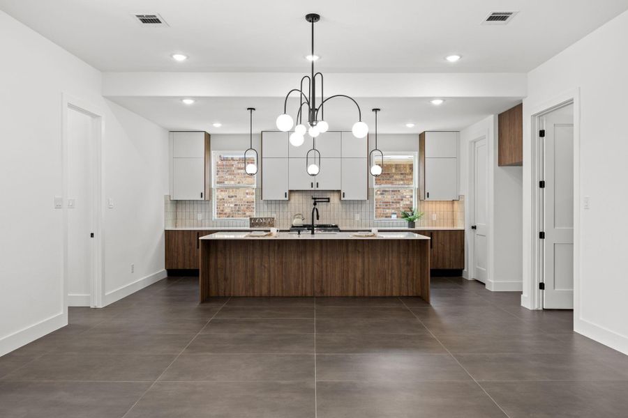 This photo showcases a modern kitchen with a large island featuring wood cabinetry and a sleek countertop. The room has white walls, contemporary pendant lighting, and tiled flooring. Ample cabinet space and a stylish backsplash complement the clean, open layout.